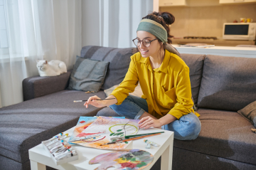 eine Frau sitzt gut gelaunt auf dem Sofa und malt ein buntes Bild 